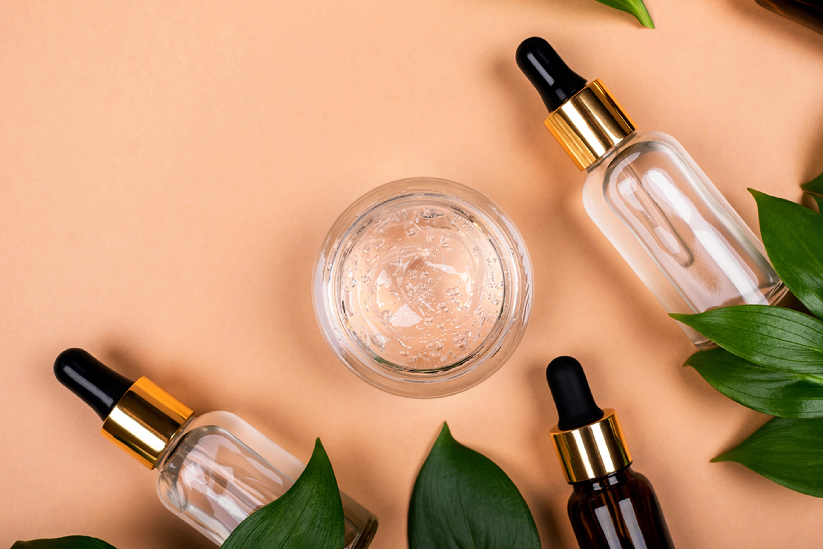 Jar of delicate clear moisturizer face gel, cream and bottles face serum on pastel beige background with green leaves.