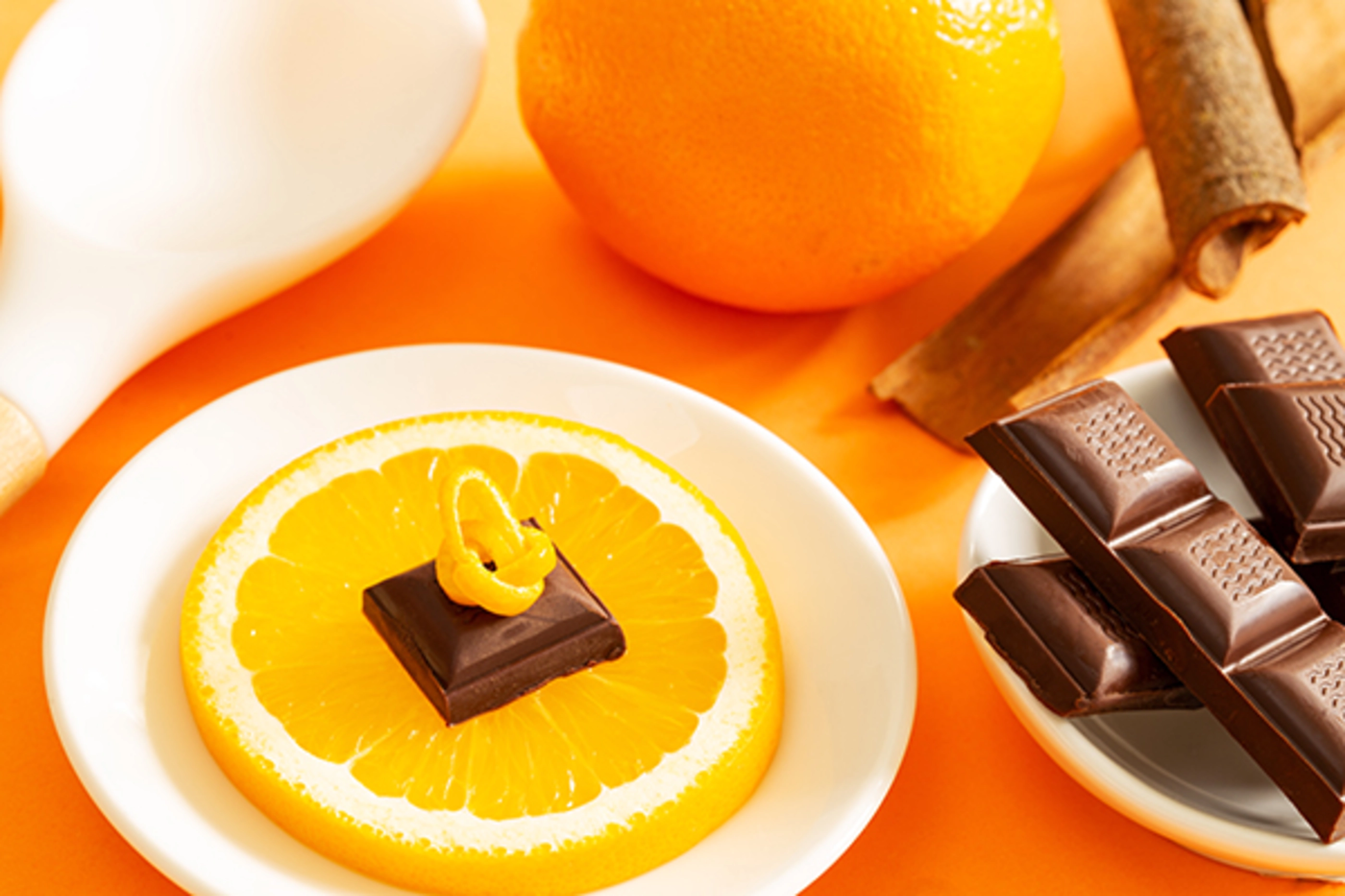 Chocolate piece with orange peel over an orange slice in a plate, chocolate bars, full orange, cinnamon sticks and white cooking spoon on the side, over an orange background.